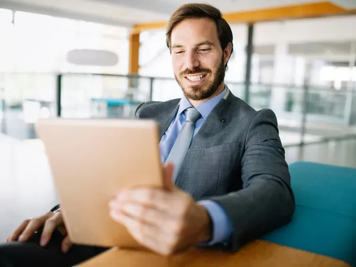 Smiling businessman in suit sitting at modern office desk holding tablet and reviewing online business content